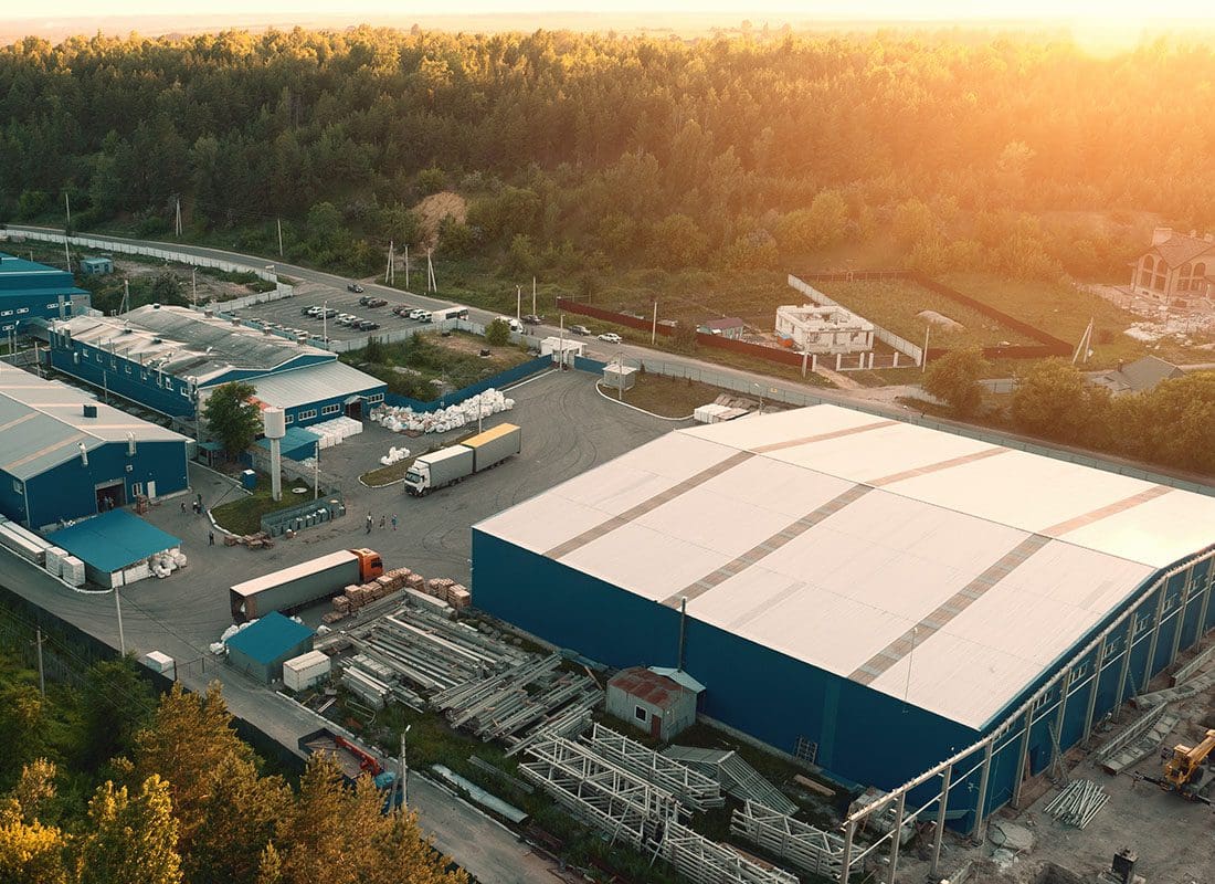 Insurance Solutions - Aerial View of an Industrial Warehouse Complex with Trucks Surrounded by Green Trees at Sunset