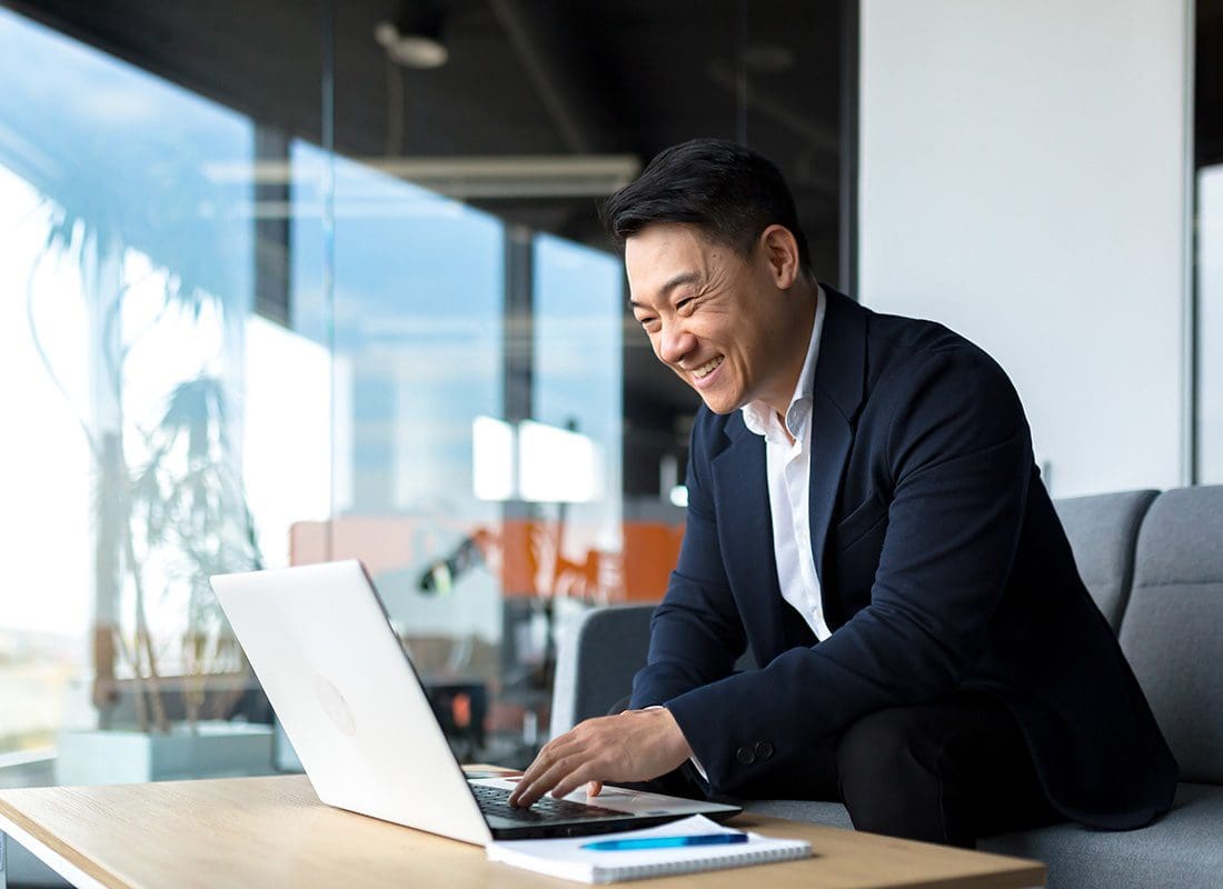 Service Center - Portrait of a Cheerful Mature Businessman in a Suit Sitting on a Chair in a Modern Office Space While Working on a Laptop
